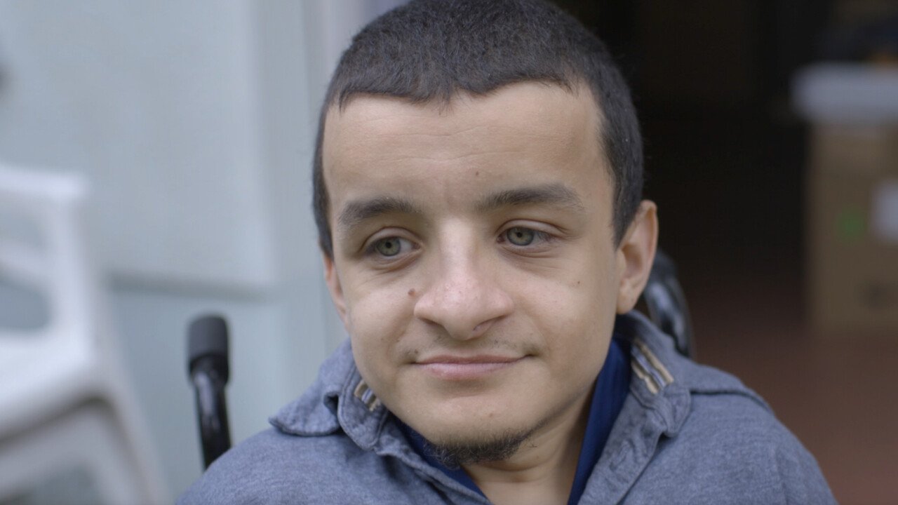 Portrait of a man with short hair and with green eyes looking slightly down. behind him there are handles of a wheelchair visible.