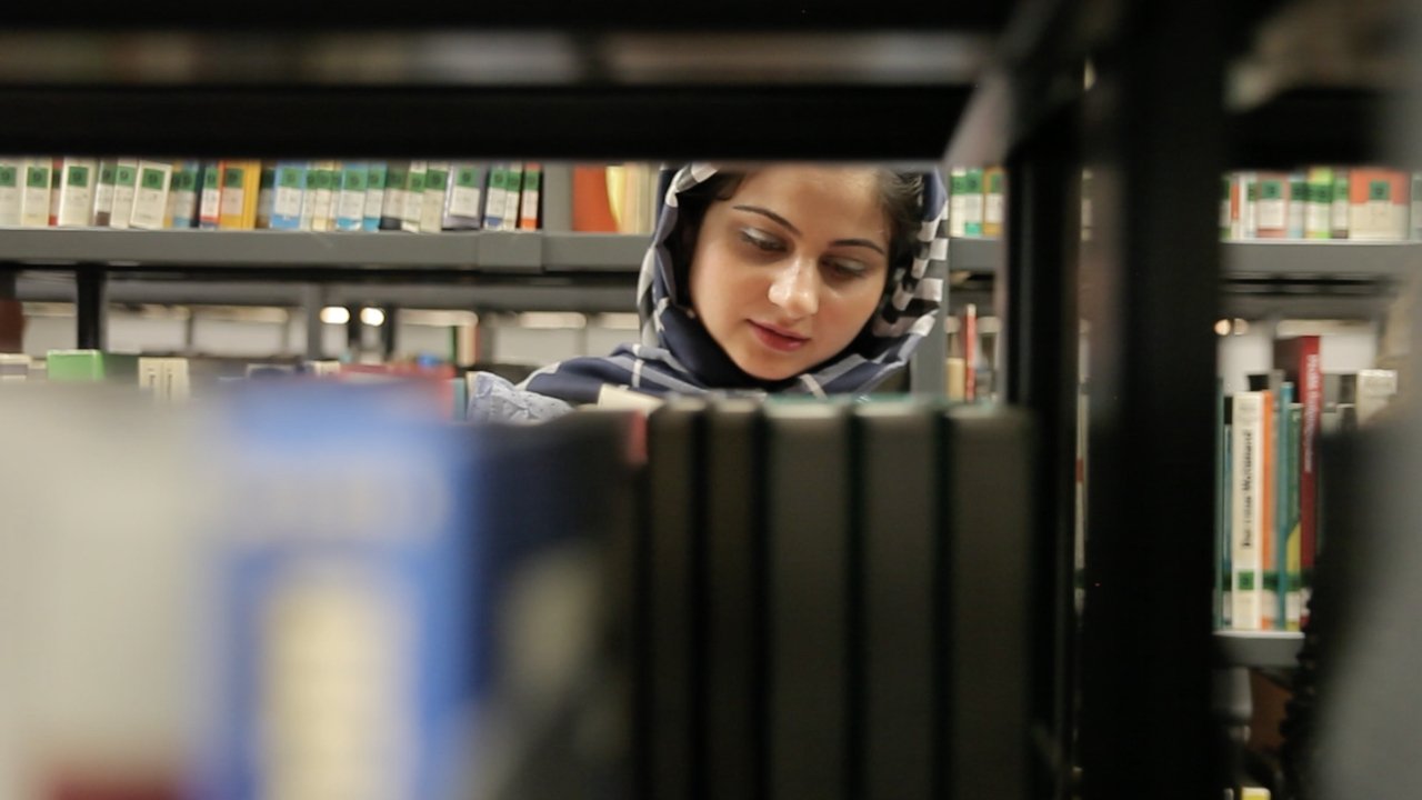 woman with headscarf looking down and reading in a library. the woman is visible through a bookshelf.
