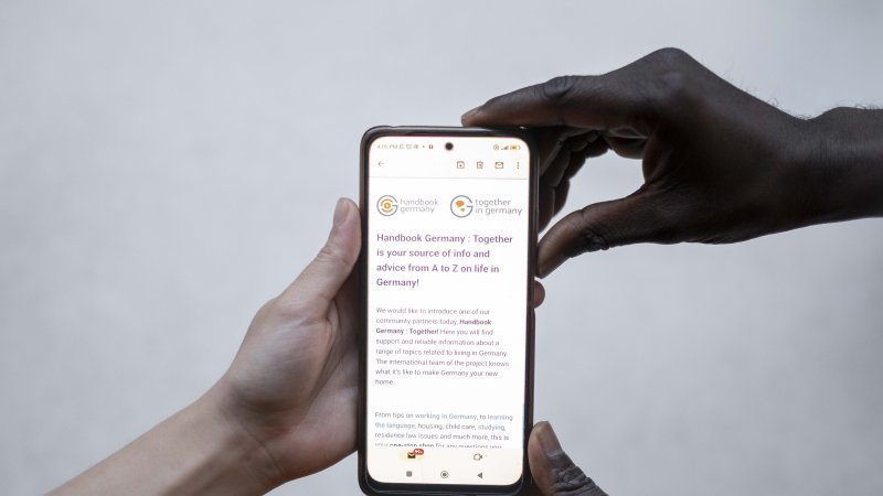 Eine weiße und eine schwarze Hand halten ein Smartphone, auf dessen Bildschirm die Logos "Handbook Germany" und "Together in Germany" zu sehen sind.