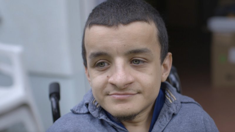 Portrait of a man with short hair and with green eyes looking slightly down. behind him there are handles of a wheelchair visible.
