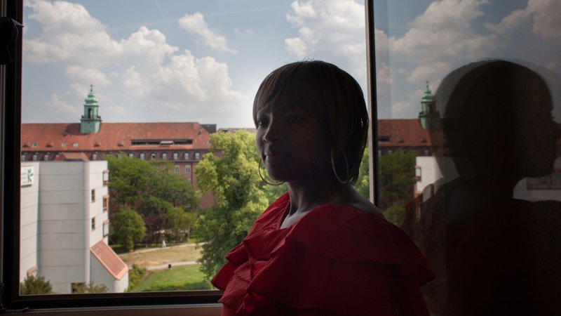 black woman standing infront of open window with her reflection in the window. background with sky, clouds, trees and a few building.